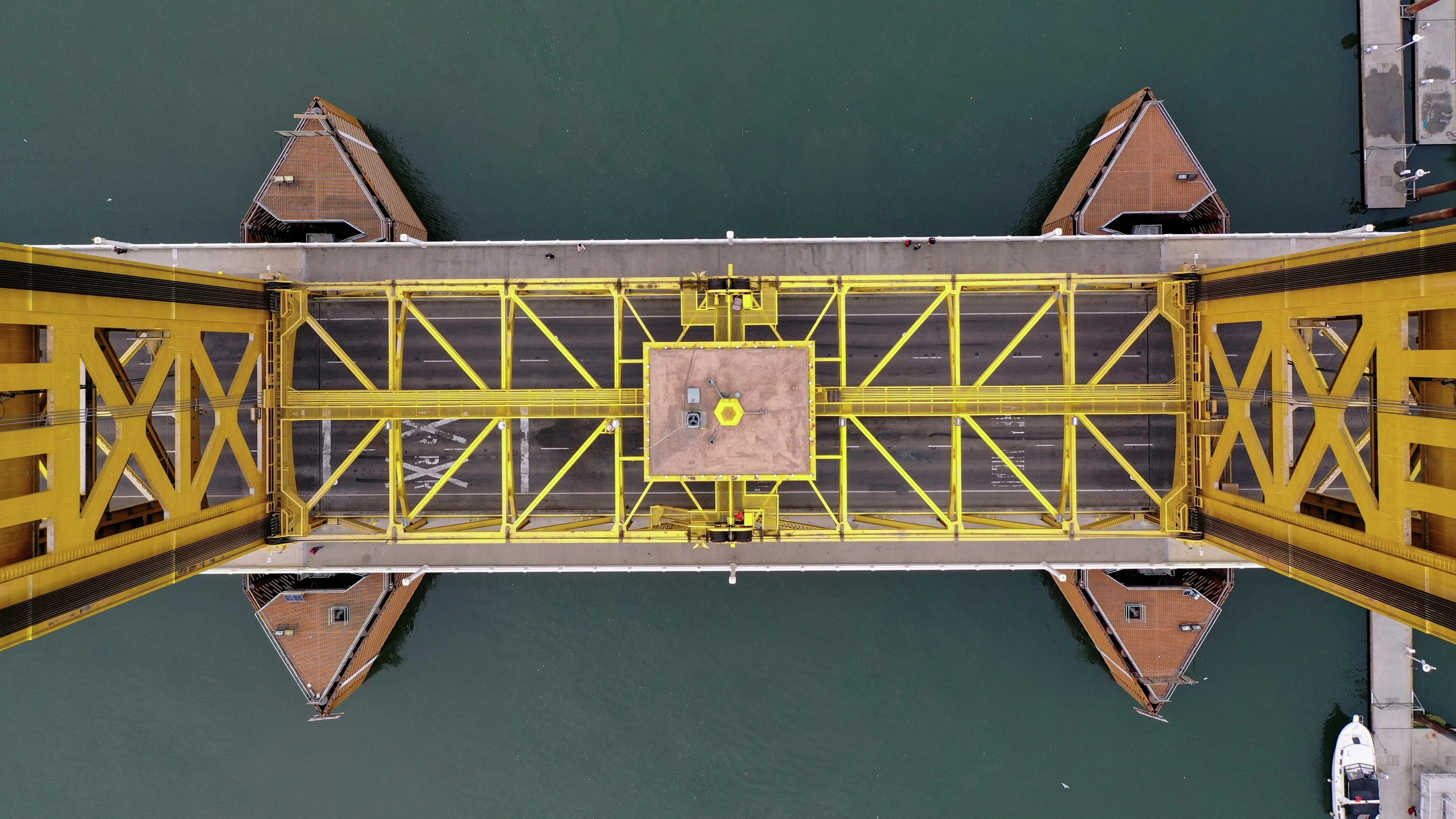 Top-down view of a yellow steel drawbridge spanning water, with symmetrical piers and road markings visible.
