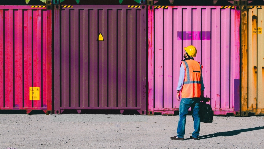 DP World port worker in safety gear standing in front of stacked, colorful cargo containers at a container terminal.