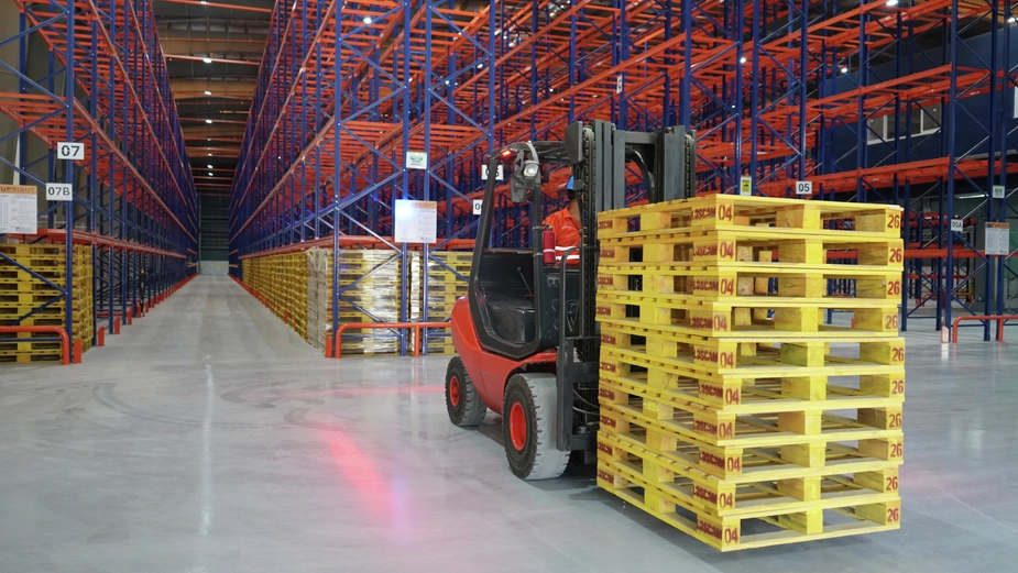 Warehouse forklift moving palletised goods along an aisle between high-density storage racks, illustrating material handling and logistics operations.