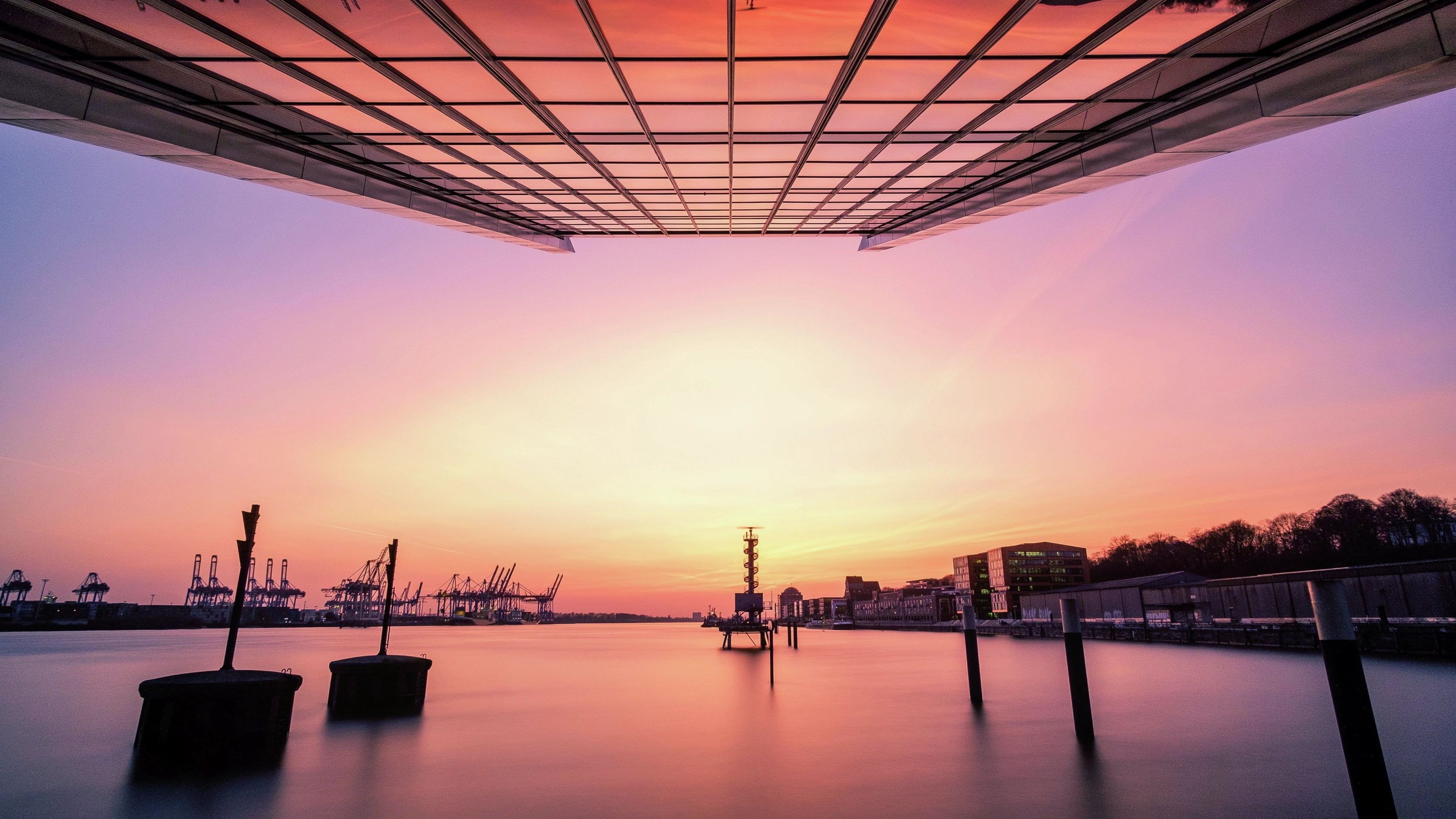 Underside of a bridge framing a calm harbor at sunset, with silhouetted cranes, docks, and city buildings reflecting on the water.